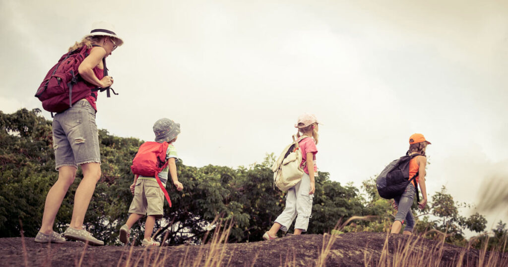 A woman is going for a hike with her 3 children.  They are all wearing summer clothes, sunhats, running shoes, and a small daypack.  They are walking on a log, one behind the other.  The mother is in the back, following the lead of her children.